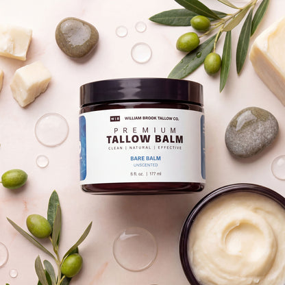 Jar of William Brook Tallow Co. Premium Tallow Balm surrounded by stones, soap bars, and olive leaves on a light background.