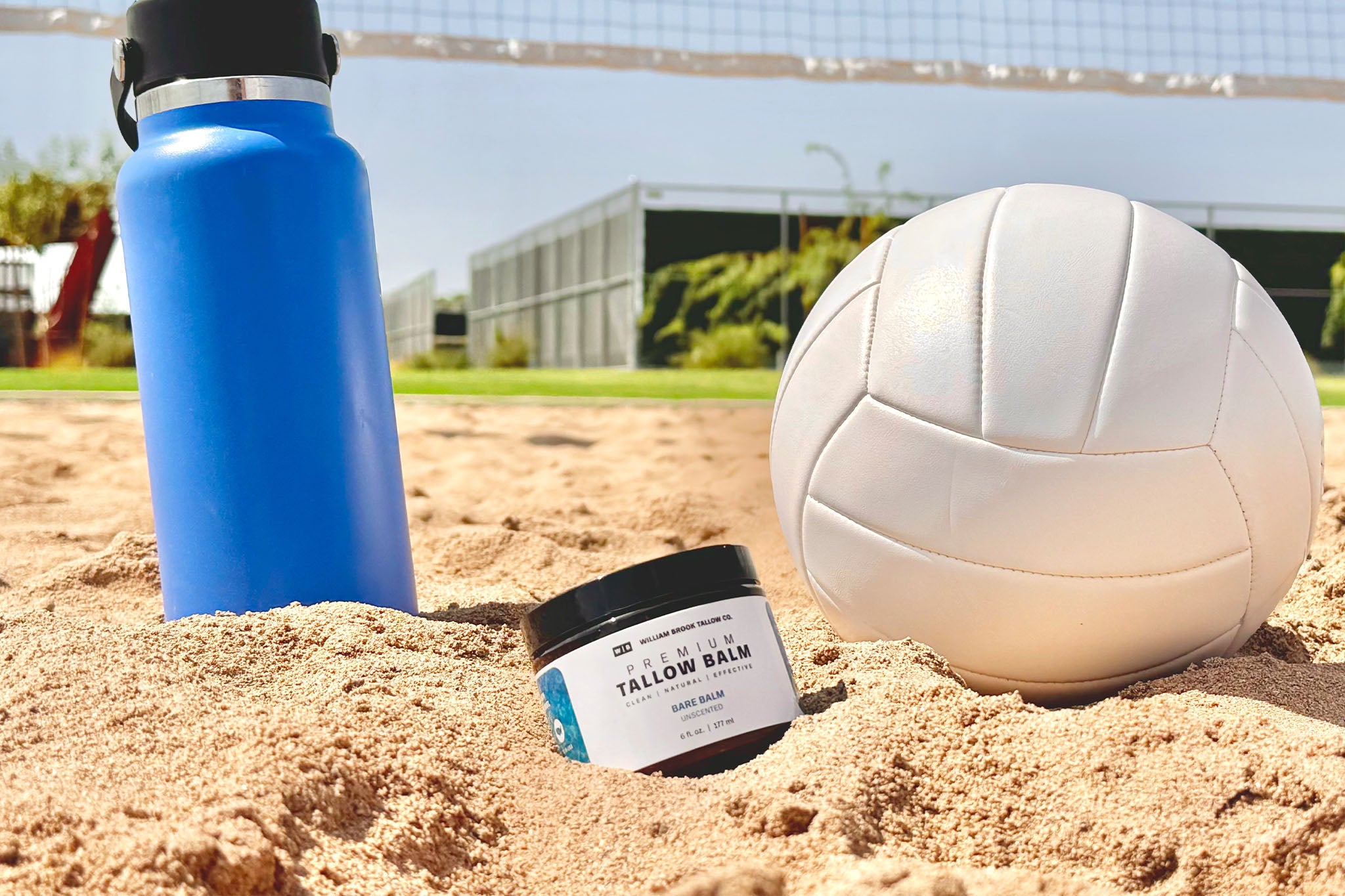 Tallow balm jar, blue water bottle, and white volleyball on sand with sports field background